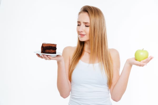 Confused cute young woman choosing between healthy and unhealthy food over white background.